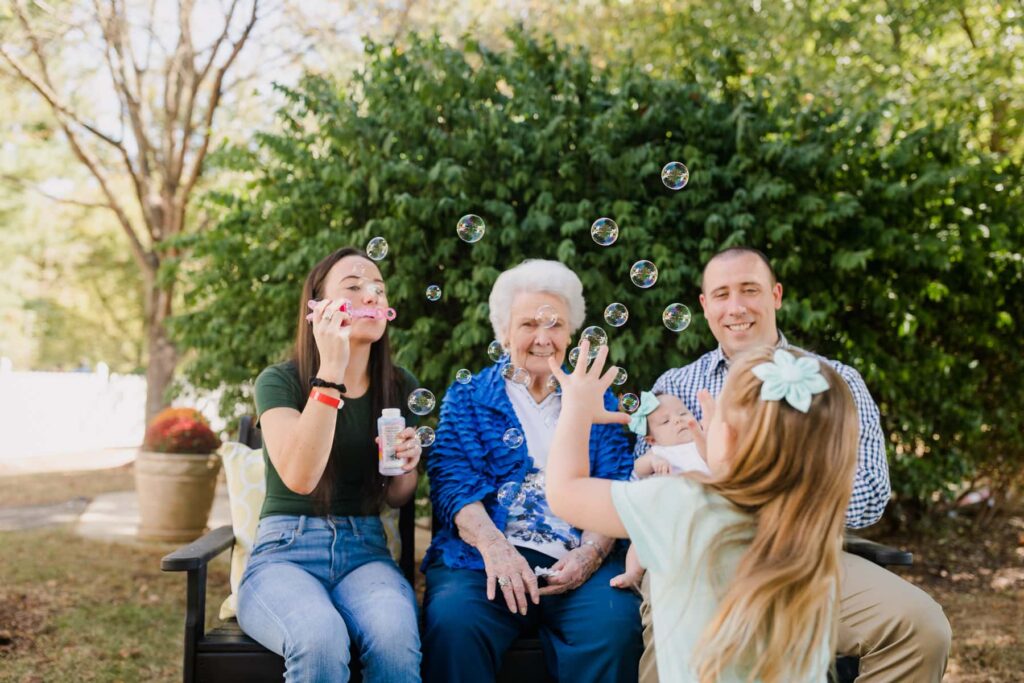 mother blowing bubbles for young daughter, sitting with husband and grandmother