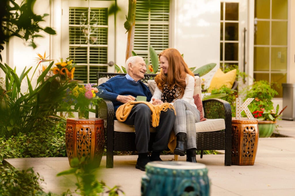 Elderly man and young woman smiling at each other on a bench, enjoying a peaceful summer moment—highlighting joyful at-home activities for dementia care.