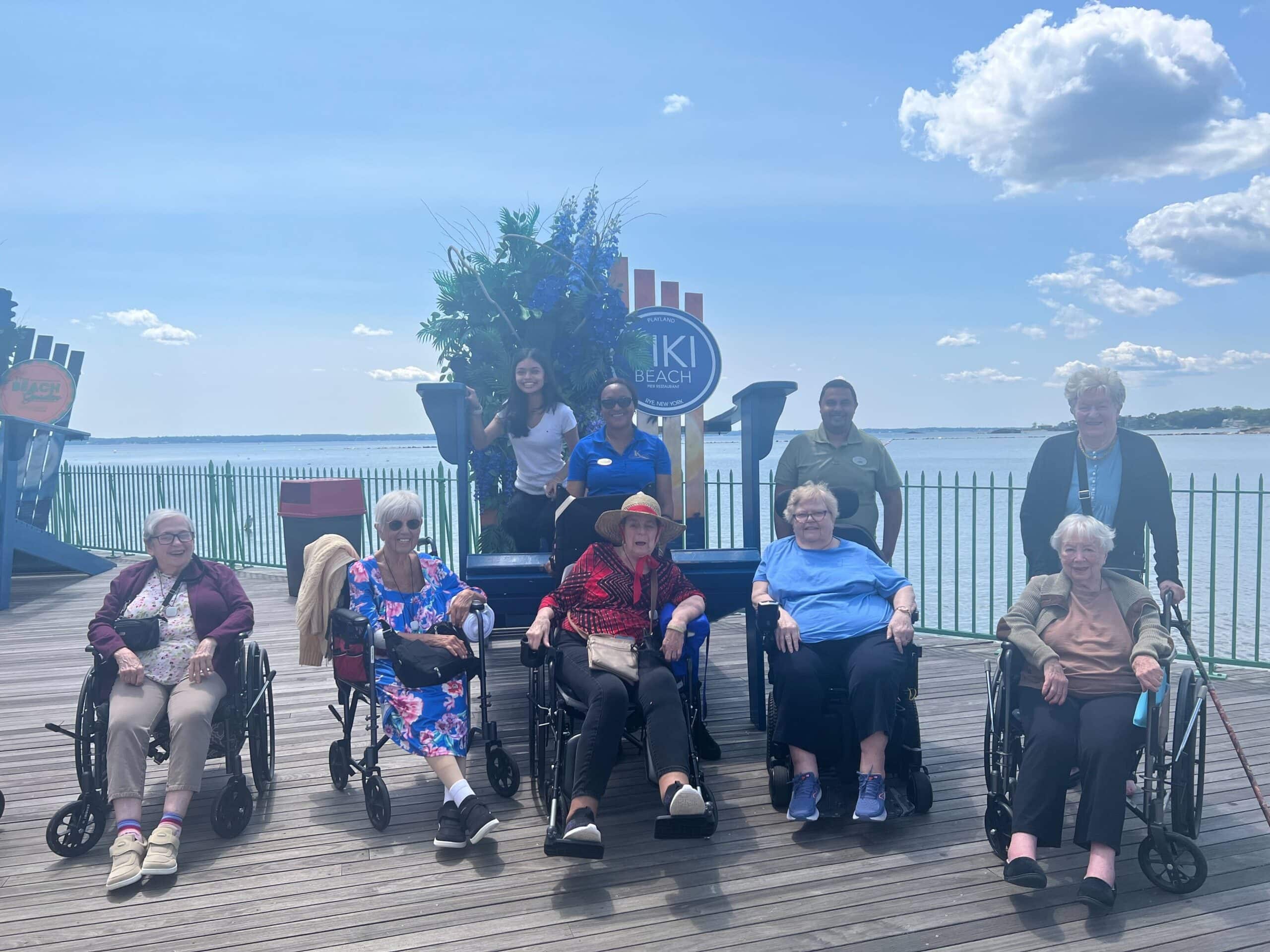 5 residents in wheelchairs on dock looking over water with caregivers staff behind them